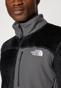 Zwarte en grijze fleece jas met een hoge kraag, gestructureerde stof, ritszak en wit logo op de borst. Patrone detail zichtbaar.