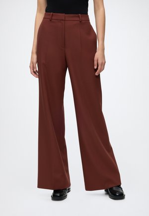 Weit geschnittene braune Hosen, getragen mit schwarzen Loafers und einem schwarzen Oberteil, von der Taille bis zu den Schuhen auf einem schlichten Hintergrund gezeigt.
