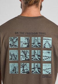 Bruine katoenen t-shirt met een grafisch ontwerp van negen blauwe geïllustreerde vierkanten die natuurtaferelen en bomen weergeven, met tekst erboven en een logo eronder.