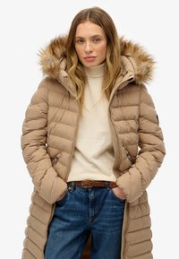 Beige pufferjakke med en aftagelig hætte i pels, horisontal quiltning og frontlynlås; båret over en lysefarvet sweater og blå jeans.