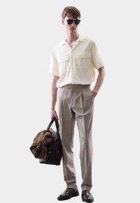 Helles, gelbes Kurzarmhemd mit zwei Brusttaschen, beigefarbene, plissierte Hose und braune Lederschuhe. Braune Tasche über einer Schulter getragen.