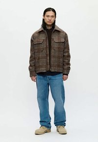 Homem de cabelo comprido, vestido com uma jaqueta de fecho castanha em padrão xadrez, suéter escuro, jeans azuis largos e botas bege, em fundo branco.