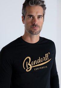 Langärmliges schwarzes T-Shirt mit goldbesticktem Text "Bendorff CONTRASTS" auf der Vorderseite. Glatte Textur, figurbetontes Design.