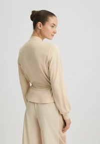 Haut beige à manches longues avec une taille ajustée, des manches bouffantes et une texture lisse. La vue arrière montre un design épuré et minimaliste avec un drapé subtil.