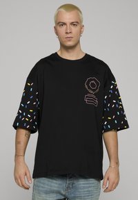 Černé oversized tričko s barevnými vzory posypu na krátkých rukávech a grafickým designem na přední straně v růžové a bílé barvě.