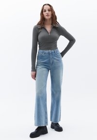 Langärmeliger, grauer Zip-Pullover, kombiniert mit hellblauen, hochtaillierten Jeans und schwarzen klobigen Stiefeln. Glatte Textur, lässige Passform und weit geschnittenes Design.