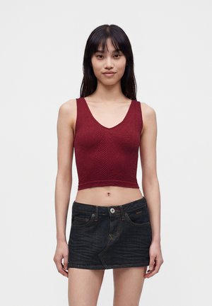 Νεαρή γυναίκα φοράει κόκκινο αμάνικο crop top με υφή μοτίβου και μαύρη μίνι φούστα από τζιν, στέκεται μπροστά από ένα απλό φωτεινό φόντο.