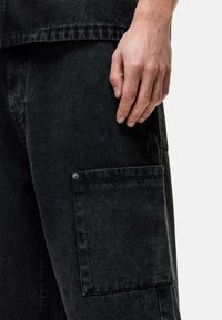 Pantalon cargo en denim noir avec une poche latérale et un détail de bouton argenté. Le tissu a une légère texture et est légèrement usé. Une main repose sur le pantalon.