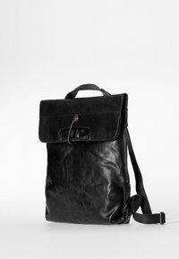 Zaino in pelle nera con pattina, manico superiore, tracolla regolabile e grande dettaglio in metallo. Texture liscia, forma rettangolare moderna.
