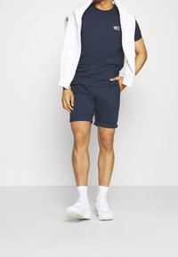 Mörkblå t-shirt med en liten logotyp, matchande shorts. En vit sweatshirt är draperad över axlarna. Modellen bär vita sneakers.