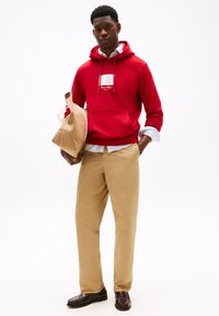 Red hoodie with a front pocket and white logo, beige chinos, and brown leather shoes, holding a tan bag. Smooth texture, casual design.