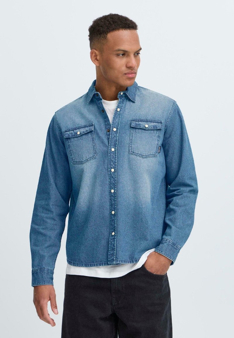 Denim shirt in light blue with long sleeves, button-up front, two chest pockets, and a casual fit. Worn over a white T-shirt.