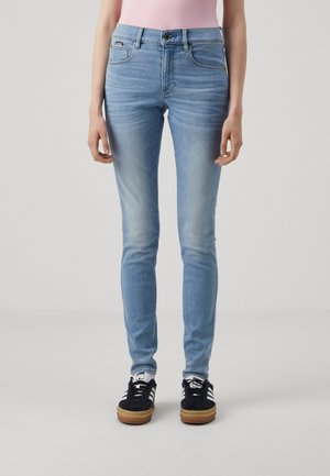 Personne portant un jean skinny bleu clair, un top rose sans manches, des chaussettes blanches et des baskets Adidas noires, debout sur un fond gris clair uni.