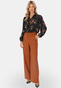 Kobieta w czarnej bluzce w kwiaty z kokardą i wysokich, szerokich, rdzawych spodniach, stoi z jedną ręką w kieszeni.