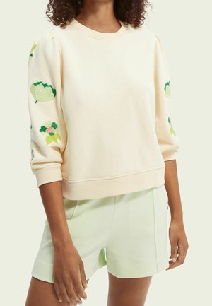 Krémová mikina s nafouknutými rukávy vyšívanými zelenými a růžovými květinovými vzory, kombinovaná se světle zelenými šortkami na stojící osobě.