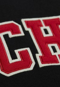 Nahaufnahme eines schwarzen Stoffes mit kräftig roten und weißen gestickten Buchstaben "C" und "H", die auf der Oberfläche aufgenäht sind.