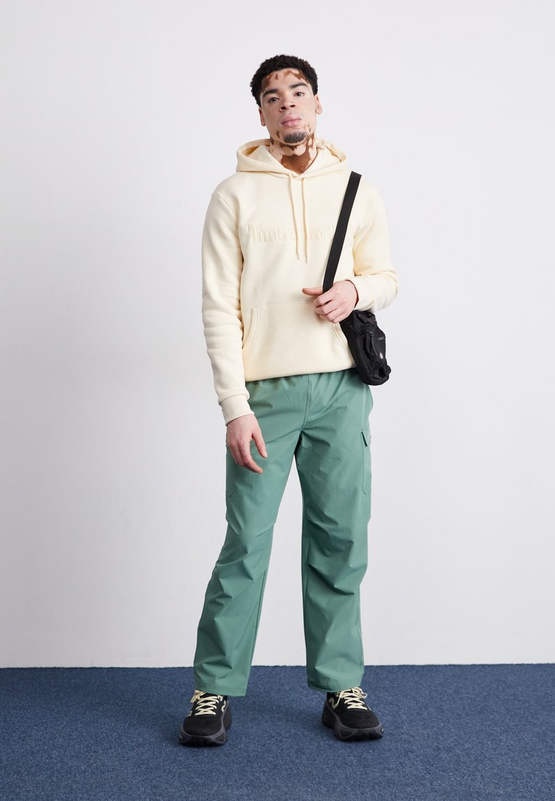 Sudadera con capucha beige con letras en relieve, combinada con pantalones cargo verdes de corte holgado y zapatillas negras. Pequeño bolso cruzado negro.