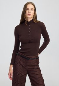 Braun gerippter Strick-Oberteil mit hohem Kragen, ausgestattet mit einer Reihe dunkler Knöpfe auf der Vorderseite. Kombiniert mit gemusterten, weit geschnittenen Hosen.