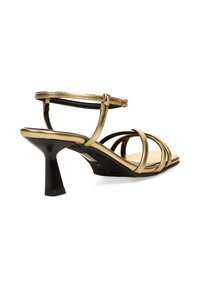 Sandalias de tacón con tiras metálicas doradas, con un estrecho tacón de aguja negro, correa para el tobillo ajustable y diseño de punta abierta. Acabado suave y brillante.