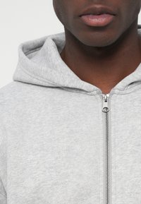 Tuvinājums uz personu, kas valkā gaiši pelēku ar rāvējslēdzēju aizvērtu kapuci, rādot tikai lūpas un apakšējo sejas daļu.