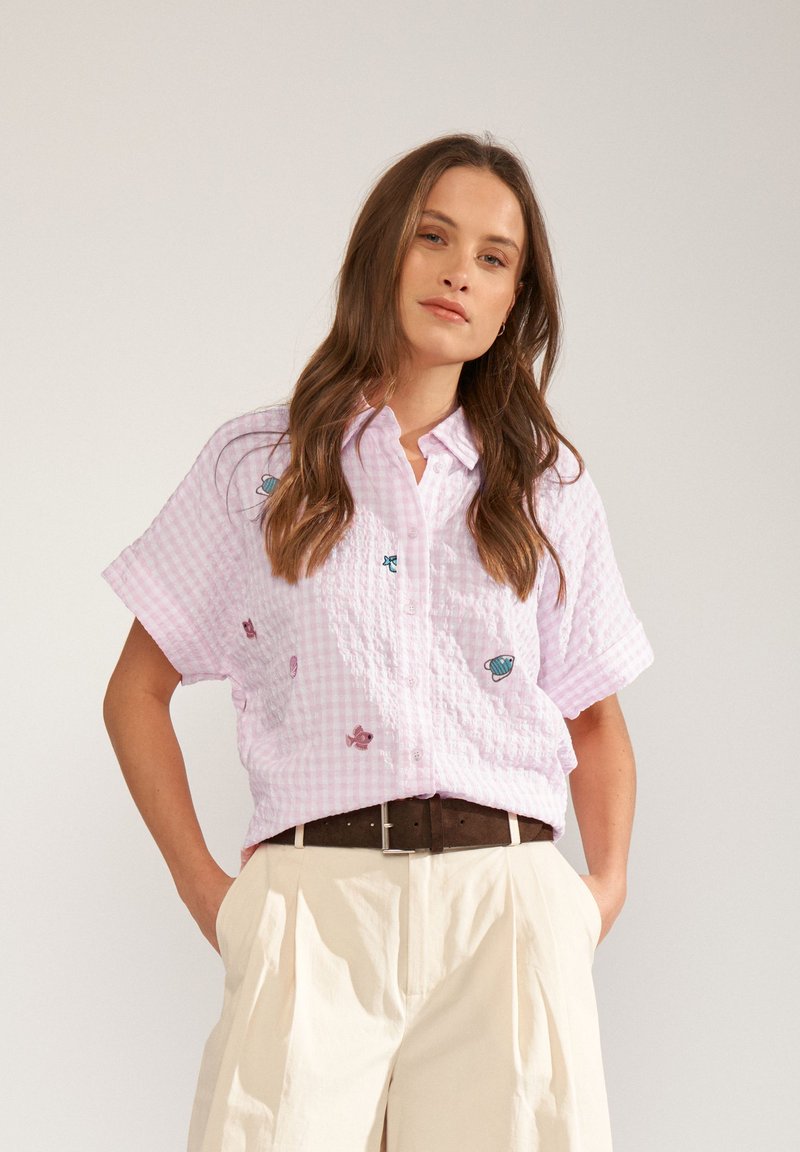 Femme portant une chemise à manches courtes à carreaux rose clair avec de petits motifs de poissons et coquillages, pantalon beige taille haute et ceinture marron foncé.