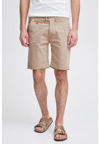Homme portant un short beige jusqu'aux genoux, un T-shirt blanc et des sandales marron clair à boucles, se tenant sur un fond blanc.