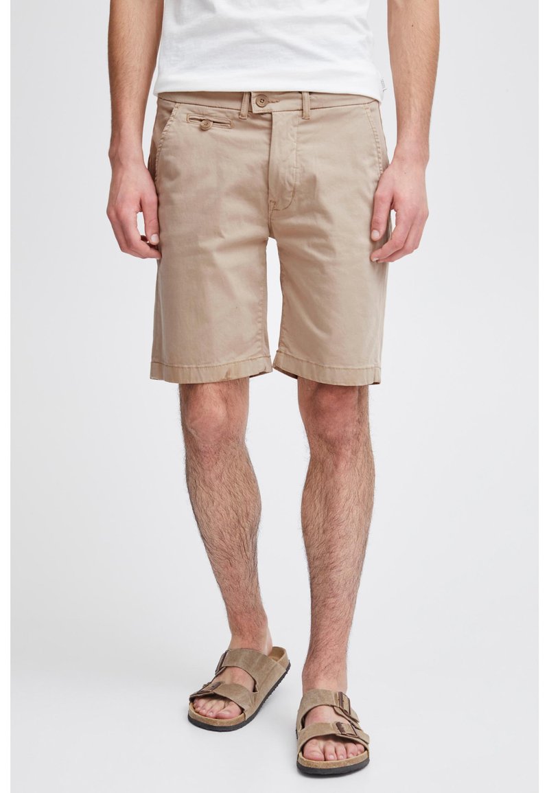 Homme portant un short beige jusqu'aux genoux, un T-shirt blanc et des sandales marron clair à boucles, se tenant sur un fond blanc.