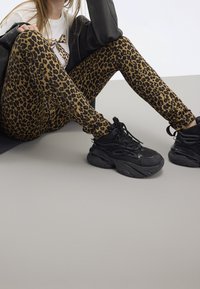 Kvinna som sitter med korslagda ben, iklädd leopardmönstrade leggings, svarta chunky sneakers, vit t-shirt och svart jacka på en grå golv.