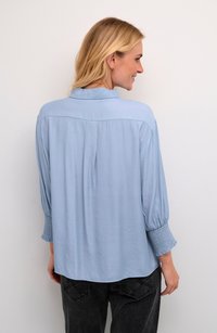 Hellbluse mit Kragen, weiten Ärmeln mit Smockbündchen und lockerer Passform. Der Stoff wirkt glatt mit einem leichten Glanz.