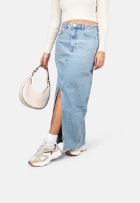 Lichtblauwe denim maxi rok met een voorplitsing, gecombineerd met witte sneakers en een beige handtas. De stof is gestructureerd en casual.
