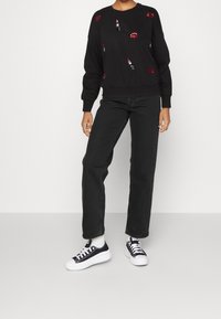 Musta huppari, jossa värikkäitä kirjailtuja huulipuna- ja suudelma-aiheisia kuvioita. Yhdistetty korkeavyötäröisiin mustiin farkkuihin ja mustiin tennareihin.