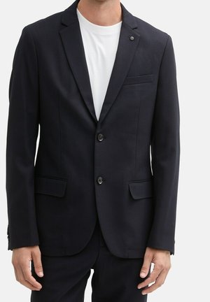 Homme portant un blazer noir à deux boutons sur un t-shirt blanc uni à col rond, debout devant un fond blanc.
