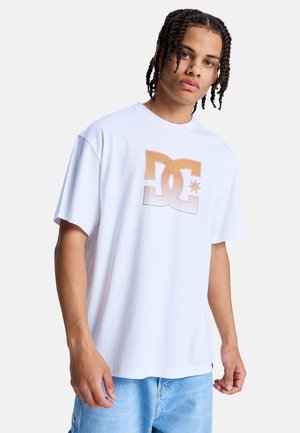Jeune homme aux cheveux tressés portant un t-shirt blanc avec un grand logo marron de DC Shoes et un jean bleu clair sur fond blanc.