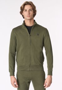 Giacca a zip verde oliva realizzata in morbido materiale, con colletto alto, polsini a coste e tasche. Dettaglio del logo discreto sul petto.