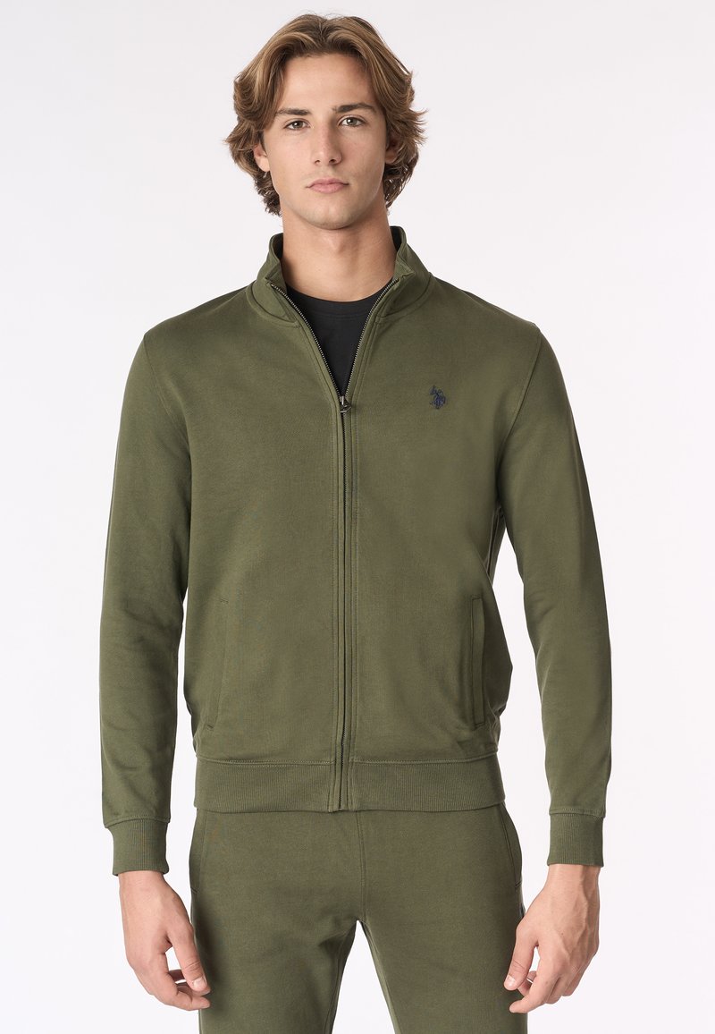 Giacca a zip verde oliva realizzata in morbido materiale, con colletto alto, polsini a coste e tasche. Dettaglio del logo discreto sul petto.