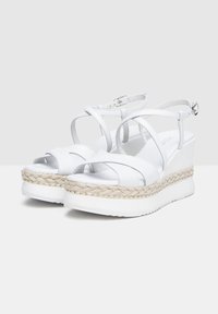Sandali con zeppa bianca e straps intrecciati, caratterizzati da una soletta in juta intrecciata e una superficie liscia, arricchiti da piccole borchie dorate.