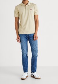 Béžové polo tričko s krátkými rukávy, zapínáním na tři knoflíky a malým červeným logem. V kombinaci s modrými džínami a bílými teniskami.