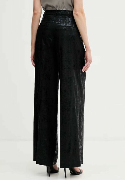 Persoon die zwarte, high-waisted, wijde fluwelen broek en zwarte hoge hak sandalen draagt, staand tegen een eenvoudige lichte achtergrond.