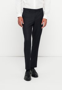 Pantalones en gris oscuro, ajustados y confeccionados en tela texturizada. Con cintura con pasadores y cremallera. Combinados con mocasines negros y una camisa blanca.