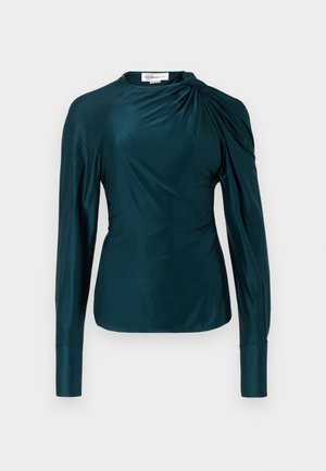 Blouse bleu sarcelle à manches longues avec détail froncé sur une épaule et poignets allongés, confectionnée dans un tissu lisse, semblable à du satin.