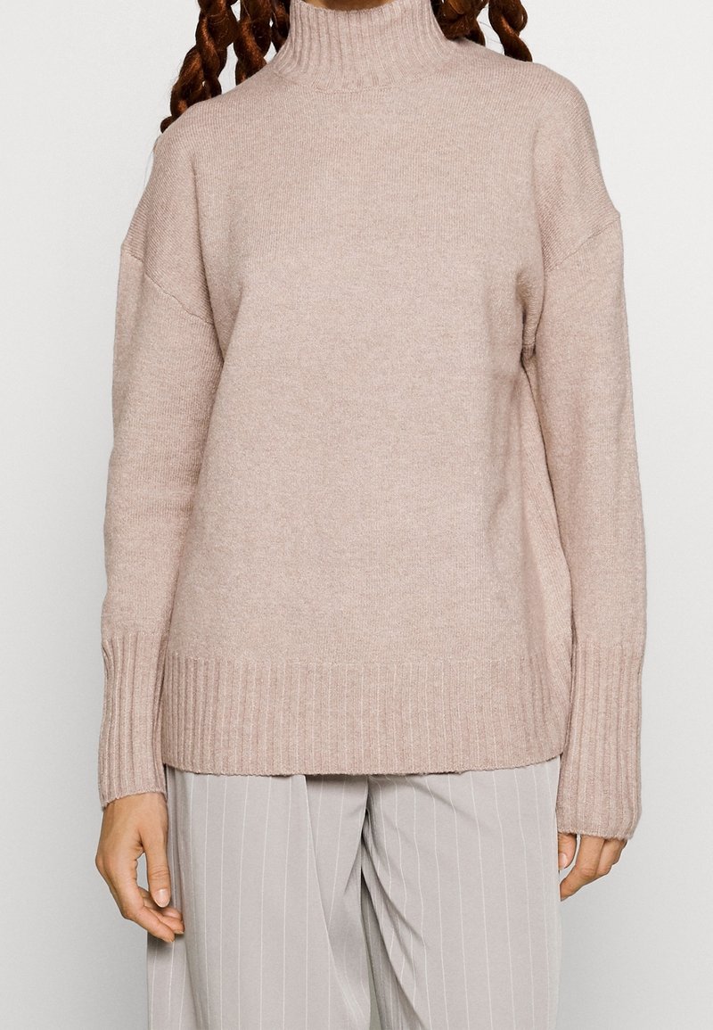 Jasno beżowy sweter z wysokim kołnierzem wykonany z miękkiego materiału, z prążkowanymi mankietami i dołem. Noszony na jasnoszarych spodni z prążkiem.