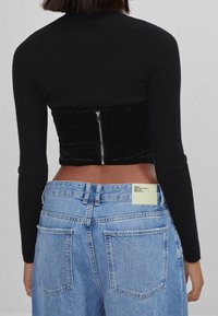 Svart ribbad långärmad topp möter en figurnära svart sammetskorsett, kombinerad med ljusblå högmidjade jeans med en etikett baktill.