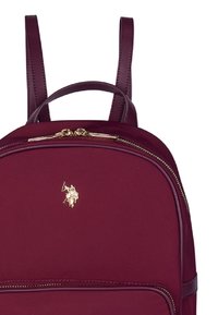 Burgundy ryggsäck med en slät tygytan, läderdetaljer, dubbla axelremmar och hardware i guldton med logotypdetalj på framsidan.