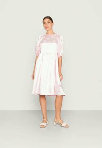 Mulher em pé a usar um vestido midi de cetim rosa claro com mangas bufantes e sandálias de salto brancas, contra um fundo liso.