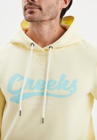 Felpa gialla con cappuccio dalla consistenza morbida, con un logo turchese "Creeks" e il testo "ORIGINAL Since 1974" sul davanti.
