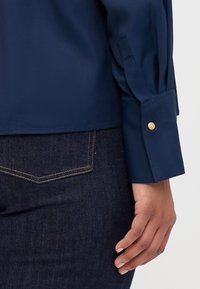 Marinblå långärmad skjorta med en slät textur, som har en knappmanchett med en metallisk guldaccent och en rak fåll, kombinerad med mörk denim.