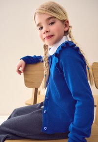 Cardigan di maglia blu con bordi arricciati, chiusura con bottoni, indossato sopra una camicia bianca con colletto. Il modello ha trecce bionde ed è seduto su una sedia.