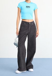 Personne portant un t-shirt cropped Nike bleu clair, un pantalon large noir, des baskets blanches, et tenant un petit sac gris dans une main.