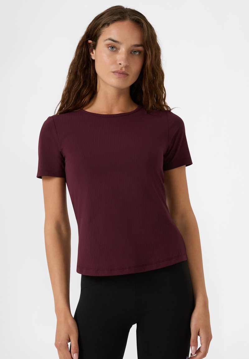 Mujer joven con cabello largo y ondulado de color marrón, vestida con una blusa ajustada de manga corta en color burdeos y pantalones negros, de pie contra un fondo blanco liso.