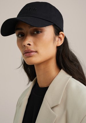 Sort baseball cap med broderet tekst, båret sammen med en off-white blazer over en sort top. Glat stof med en buet skygge.